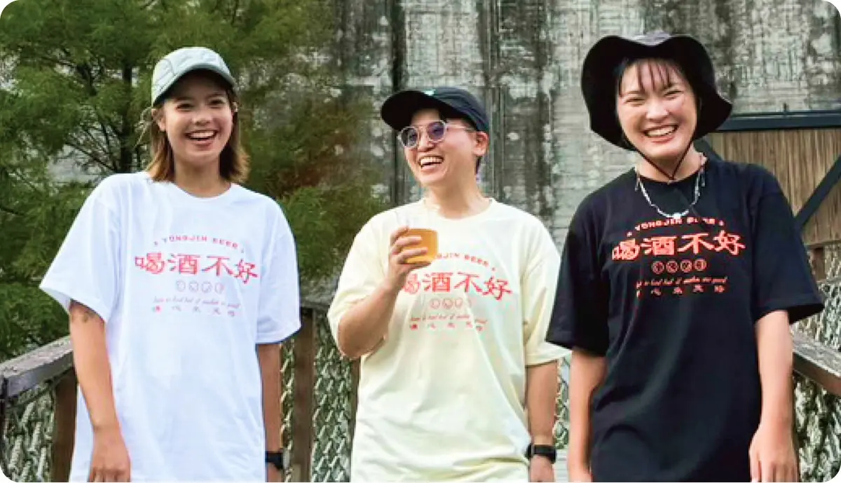 三位年輕人穿著湧金「喝酒不好」品牌 T 恤，在戶外木橋上開心合影